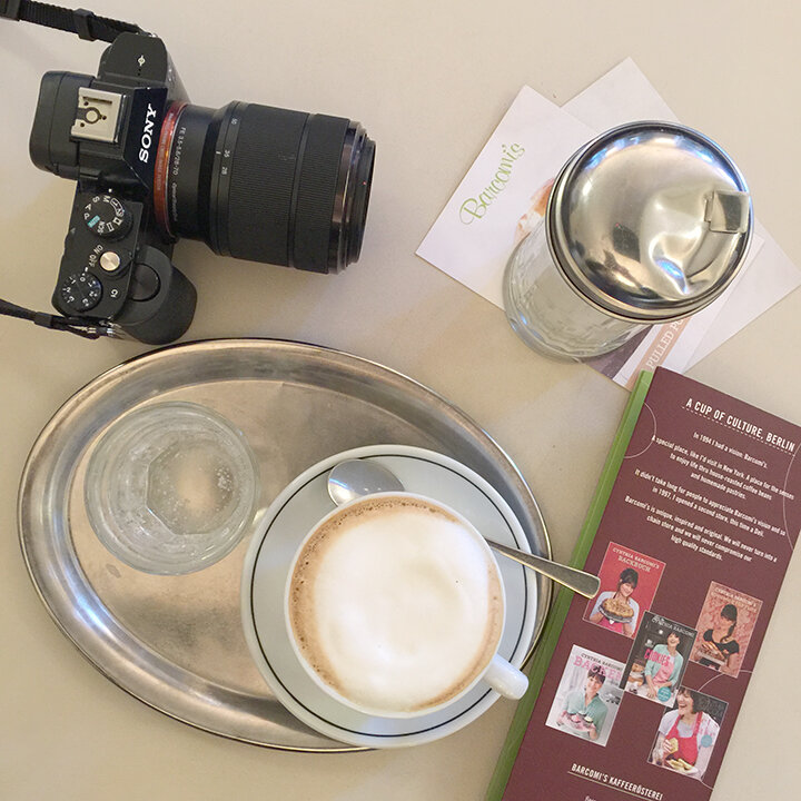 solo travel essential: camera on table with travel brochure. Cappuccino and sugar and glass of water. 
