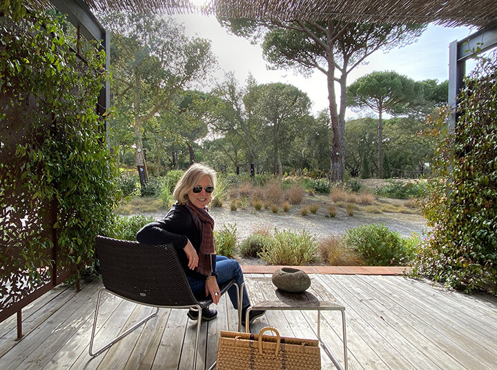 woman sitting on a chair on a terrace facing a garden. The woman is looking back at the camera. Comporta