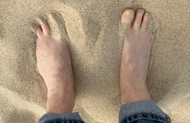two feet in sand, part of blue jeans is visible