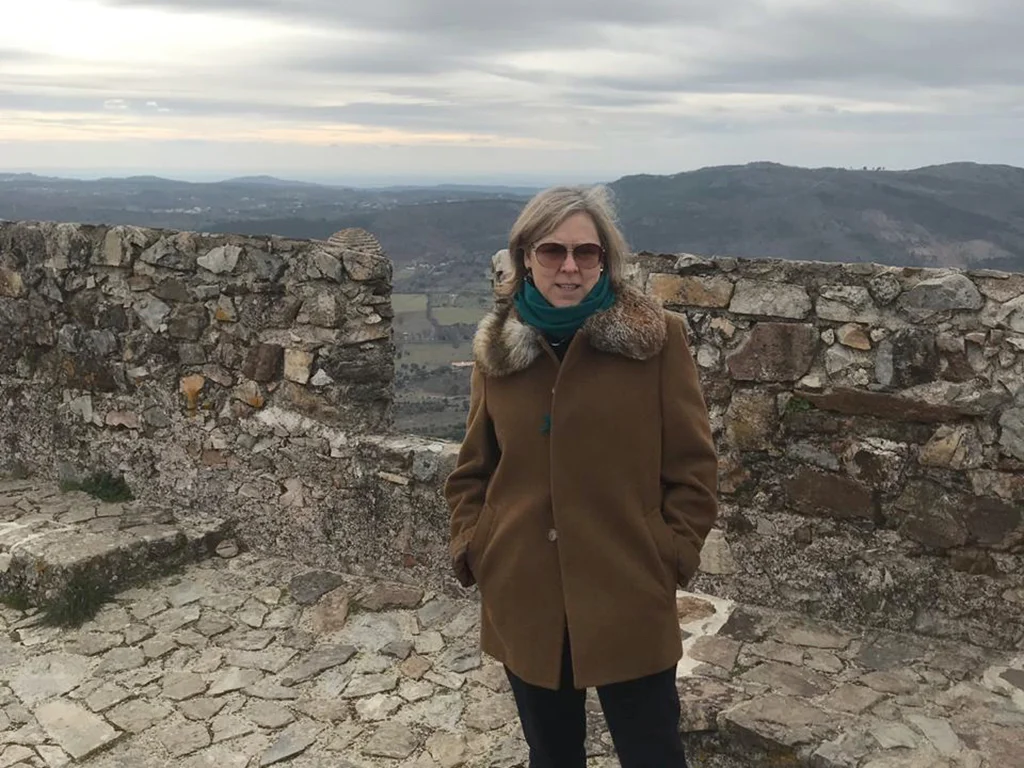 Woman wearing a typical Alentejo winter coat, details of castle wall in the background, dark clouds, hills in the background. 50 and Rising
