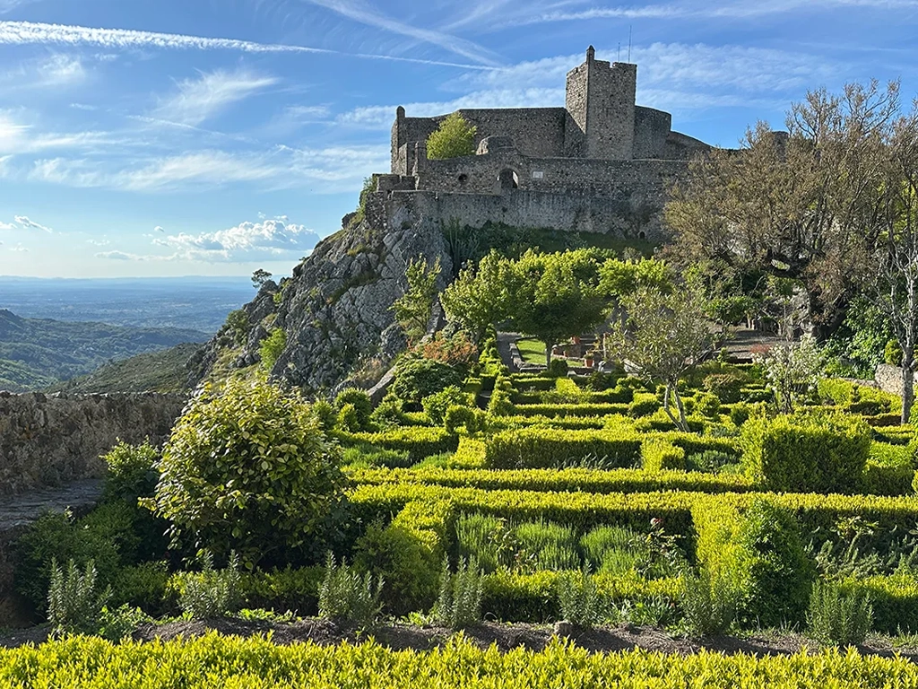 Marvao Castle on top of a hill, bright green manicured garden in the foreground. Blue sky, hills to the side. 