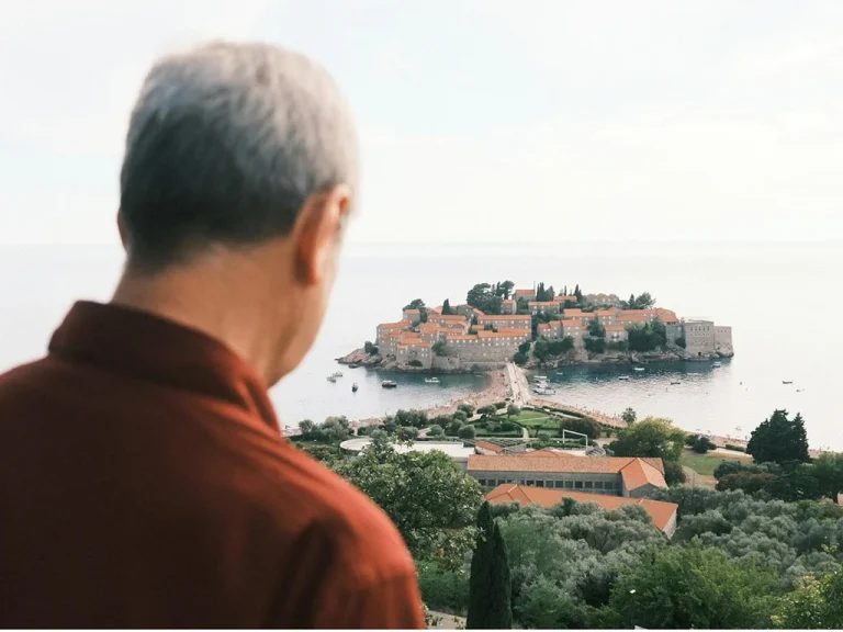 man looking at a view of an island, he is up a hill, greenery, he is looking away and wearing a red shirt, traveling over 50