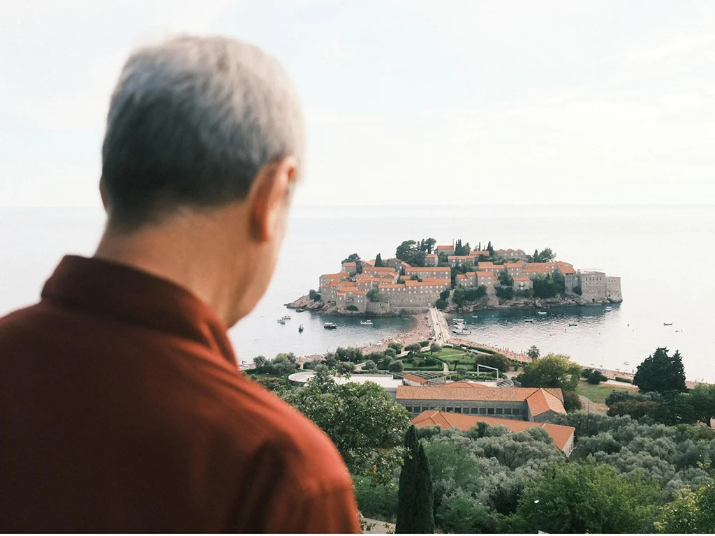 man looking at a view of an island, he is up a hill, greenery, he is looking away and wearing a red shirt, traveling over 50